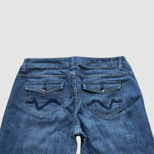 Lågmidjade Vintage Bootcut Jeans  - VARAN KAN KÖPAS DIREKT PÅ VÅR HEMSIDA, ettmarke.se MODELL: Lågmidjade & Bootcut. FÄRG: Mörkblå. STORLEK: W32 L34 enligt lapp. Uppskattad storlek 42. MIDJA RAKT ÖVER: 44 cm. MIDJEHÖJD: 23 cm. TOTALLÄNGD (FRÅN MIDJAN): 107 cm. BENLÄNGD (innerlår): 84cm