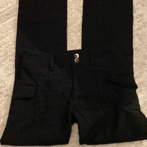 Svarta byxor - Svarta ”bootcut” byxor i tunnare material (skönare material) från Shein i storlek 32 / XS (S) som har små fickor på sidan och detaljer som två stycken knappar och ett streck i mitten ner till byxorna. 