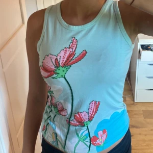 Blommig ärmlös topp från Yessica - Säljer en superfin ärmlös topp i storlek S. Toppen är ljusblå med ett färgglatt blommönster i rosa, grönt och vitt. Perfekt för sommaren och kan stylas både till vardags och fest. Materialet känns som bomull och är väldigt bekvämt att ha på sig. Passar perfekt till jeans eller shorts!