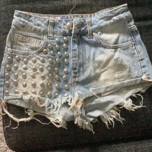 Coola jeansshorts med nitar - Säljer ett par riktigt coola jeansshorts med hög midja. De är ljusblå och har nitar på ena sidan fram. Shortsen är slitna och har fransiga kanter, vilket ger dem en grunge-look. Perfekta för sommarens alla festivaler eller bara en chill dag på stan. De är i nyskick och passar perfekt för dig som vill sticka ut!