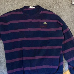 Randig tröja från Lacoste - Säljer en snygg och bekväm randig tröja från Lacoste. Tröjan är mörkblå med lila ränder och har en broderad Lacoste-logga på bröstet. Perfekt för höst och vinter, och passar både till vardags och lite finare tillfällen. Tröjan är i bra skick och väldigt skön att ha på sig.