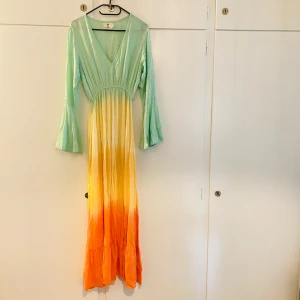 Flerfärgad långklänning från Wild Collection - Säljer en superfin långklänning från Wild Collection. Klänningen har en vacker färgövergång från mintgrön upptill till gult och orange nedtill. Den har långa ärmar och en v-ringad hals. Perfekt för sommarens fester eller en dag på stranden. Materialet känns lätt och luftigt, vilket gör den bekväm att bära hela dagen.