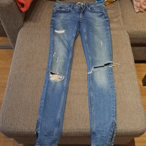 Blå slitna jeans med dragkedjor - Säljer ett par blå jeans med slitningar och dragkedjor vid bensluten. Jeansen har en tight passform och är i en klassisk blå färg. Perfekta för en avslappnad och trendig look! knappt använd. passar Xs oxh S