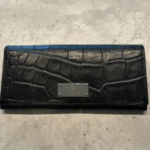 Svart plånbok från Tiger - Säljer en snygg svart plånbok från Tiger. Den är gjord av skinn med ett krokodilmönster och har en metallplatta med Tiger-loggan på framsidan. Plånboken har flera fack för kort och en genomskinlig ficka för ID-kort. Perfekt för att hålla ordning på dina kort och kontanter!