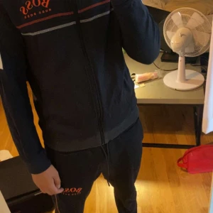 Svart hoodie från Hugo Boss - Säljer en svart hoodie från Hugo Boss. Den har en dragkedja framtill och är långärmad. Det finns en röd Hugo Boss-logga på bröstet och en grå rand ovanför loggan. Perfekt för höst och vinter. Skön och stilren, passar både till vardags och träning.