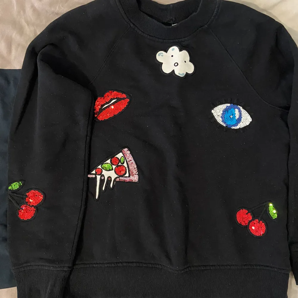 Säljer en svart sweatshirt från H&M Divided i storlek S. Tröjan har coola broderade detaljer som ett par röda läppar, en pizza slice, ett blått öga, körsbär och en söt liten molnfigur. Perfekt för en avslappnad och trendig look! 🖤🍒👁️🍕. Hoodies.