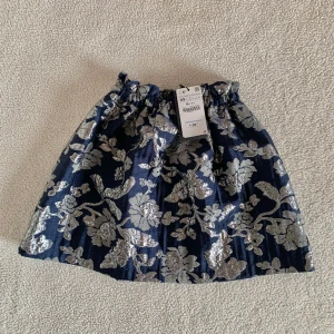 ZARA marinblå kjol med blommor - Otroligt stilig marinblå kjol från ZARA med silvermetalliska detaljer. Väldigt generös i storlek då midjan är stretchig och kjolen i en rymlig modell, passar alltså allt mellan XS-M. Ny med tags, utan anmärkningar.