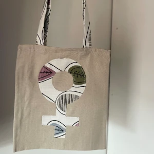 Tygkasse, tygväska, tygpåse  - Handsydd tygkasse med motivet ”feminist”.   Tillverkad i 100% återvunna textilier.   Mått: 32x35 cm  Hämtas på Södermalm alternativt skickas med post. 