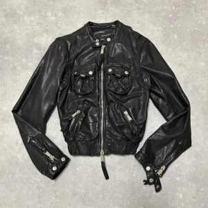 Dsquared2 skinnjacka - Dsquared2 jacka gjort av äkta skinn. Storlek IT44 men passar tajt, passar mig som har XS. Mått: längd 56 cm, axlar 45 cm, armar 63 cm, bröst 50 cm, midja 34 cm. Har många cargo fickor där framme och double zip. 