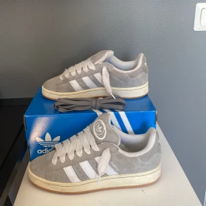 Gråa Adidas Campus sneakers - Ett par gråa Adidas Campus sneakers med vita detaljer och snörning. Skorna har en vit sula och är i bra skick.