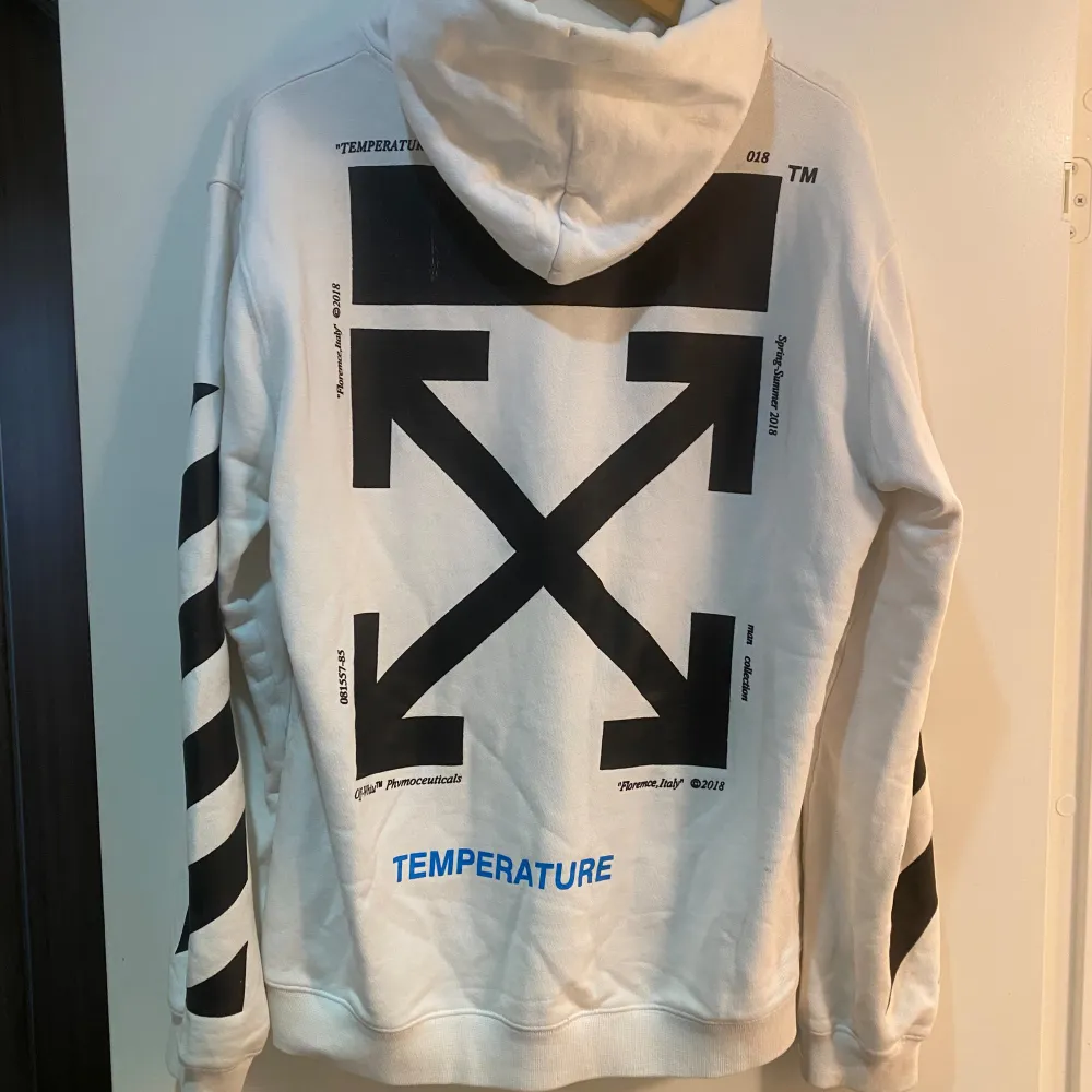 En vit hoodie från Off-White med ett stort Mona Lisa-tryck på framsidan och texten 'TEMPERATURE' i blått på fickan. Baksidan har ett stort svart grafiskt tryck med pilar och text. Ärmarna har svarta diagonala ränder. Hoodien passar som en L. Skick 7/10. Hoodies.