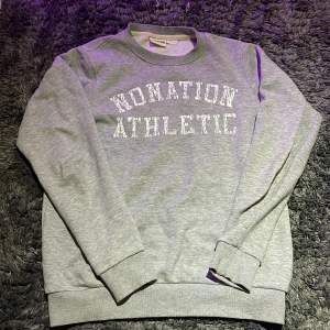 Sweatshirt från märket ”Nonation athletic” i färgen grå och ett tryck av märket ”Nonation athletic” i färgen vit. Inget slitage och inget tecken på användning.