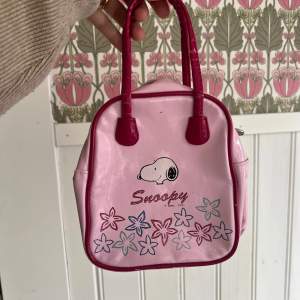 En liten rosa handväska med motiv av Snoopy och blommor. Väskan har två handtag och en dragkedja för att stänga den. Den är dekorerad med rosa och lila blommor samt texten 'Snoopy' på framsidan.