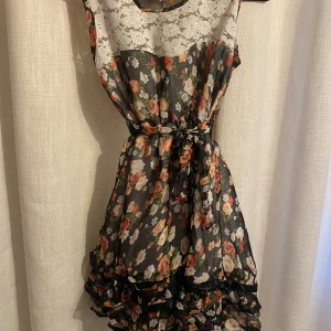 Blommig klänning - En söt, blommig lite kortare klänning men söta detaljer. Men fina volanger längst ner och vit, blommig spets högst upp. Endast använd en gång på min skolavslutning förra sommarn. Den har en liten defekt vid axeln men syns knappt. 💕