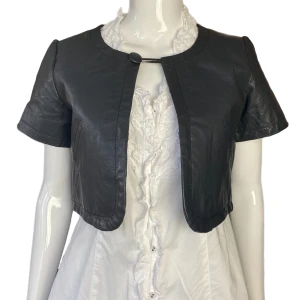 Bolero  - Bolero i fint skick i 100% skinn. Från Pieces, storlek M. Dockan använder storlek XS. Upplever färgen som svag svart  MÅTT: Axel till axel 37cm Ärmlängd 18cm  Armhåla till armhåla 47cm Längd 40cm 