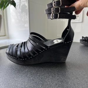 Skor i trä och läder från Swedish Hasbeens, svarta, strl 37 - Svarta ”träskor” med ovandel i läder från Swedish Hasbeens från deras samarbete med H & M. Kilklack och tre remmar kring vristen med spännen. Fint skick.