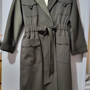 Trenchcoat - Helt ny och oanvänd. Bra kvalitet från Turkiet. Smutsig grön och perfekt för hösten och våren. Vattentät. Storlek 42.
