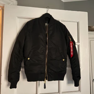 Bomberjacka Alpa Industries - Svart bomberjacka med guldiga detaljer från Alpha Industries. Ganska vadderad, perfekt till våren :) för liten i armarna på mig så får tyvärr inte på mig den. Använd nån enstaka gång för längesen! 