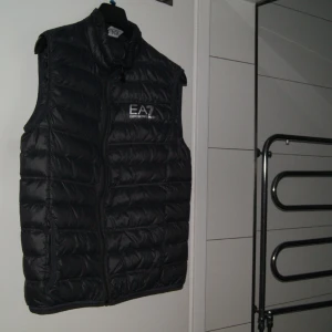 Emporio Armani - väst - Västen är i nyskick. Den har storleken S (L i barnstorlek), den är  stretchig och passar därför även till storlek-M. Modellen på bilden är 180 cm.  Den kan bäras över skjorta eller träningströja (bild 2,3). Vi har även etiketten med ursprung, material...