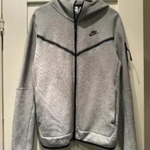 Nike Luvtröja NSW Tech Fleece - Grå/Svart - Helt ny - använd ett fåtal gånger.  Kvitto finns.   Den här huvtröjan med hel dragkedja är en mix av två av Nikes mest ikoniska looks – den tidlösa jackan Windrunner och jackan Tech Fleece.