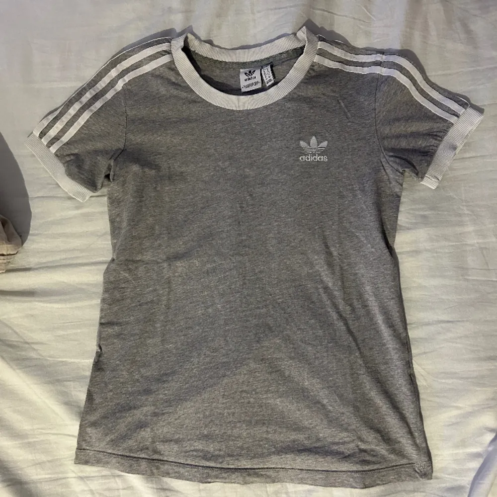 Grå t-shirt med streck över axlarna från Adidas. Fint skick, nästan oanvänd! Säljer då jag rensar ut mina kläder:). T-shirts.