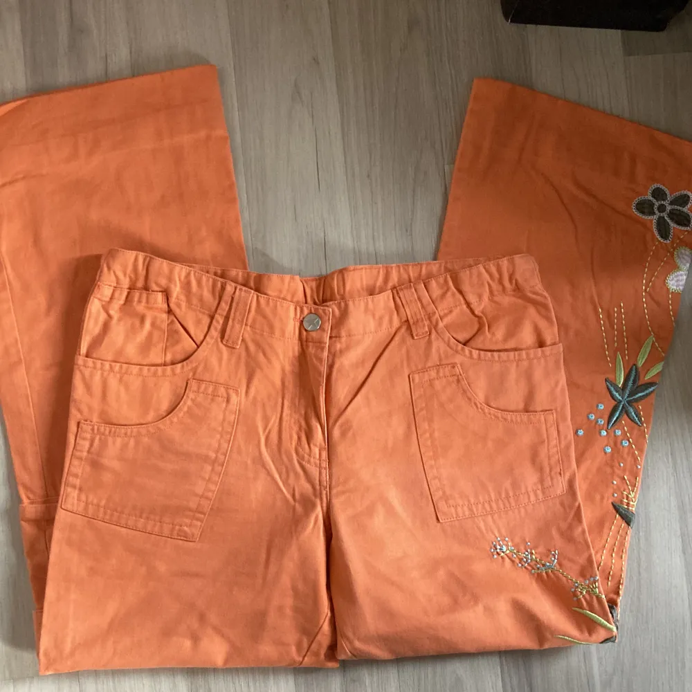Jättefina lågmidjade orange byxor med blommor på sidan av benen och en ficka. Verkligen jättefina detaljer!🧡. Jeans & Byxor.