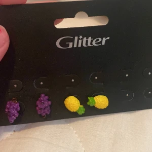 Örhängen från Glitter - Ett set med örhängen från Glitter. Setet innehåller örhängen i form av vindruvor och ananas. Vindruvorna är lila och ananaserna är gula med gröna blad.