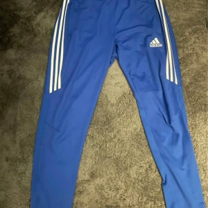 Blå träningsbyxor från Adidas - Blå träningsbyxor från Adidas med vita ränder längs sidorna och Adidas-logotyp på ena benet. Byxorna är tillverkade i ett lätt och bekvämt material.