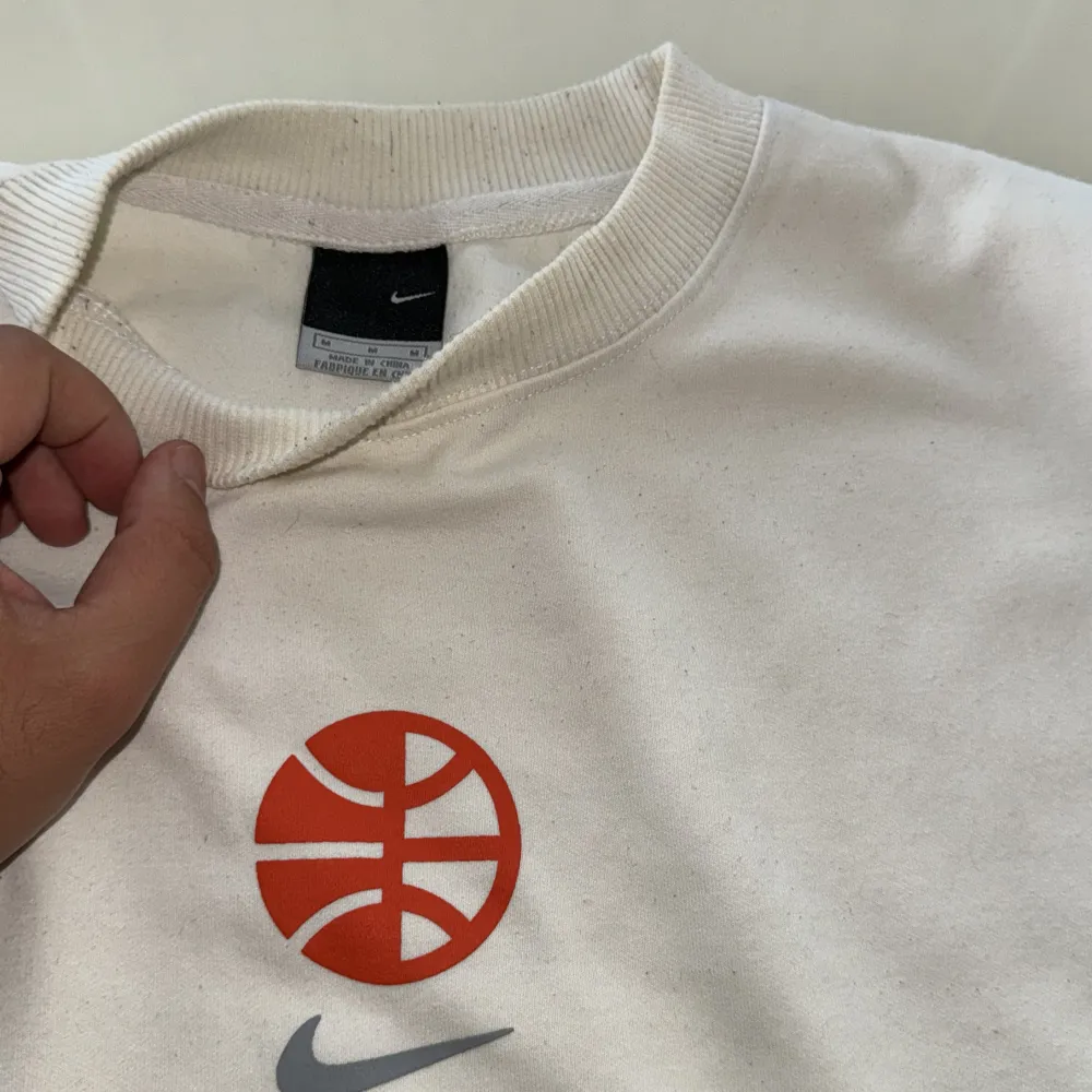 En vit sweatshirt från Nike med en röd basketboll och grå Nike-logotyp på bröstet. Tröjan har långa ärmar och ribbstickade muddar vid ärmslut och nederkant. Det finns även en etikett med texten 'Nike Hoops' på framsidan.. Tröjor & Koftor.