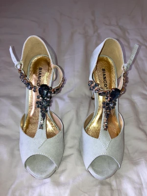 Högklackat, BIANCO - Högklackade skor från BIANCO. Ljusgrå med mörkgrå strass och guld detaljer. Strl 38 Aldrig använda! Klackhöjd ca 10cm