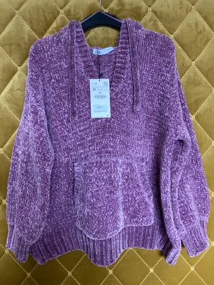 Stickad tröja ZARA - Stickad tröja från ZARA med luva. 100% polyester. Aldrig använda, prislapp sitter kvar. Strl M. Färgen är ljus syrenlila, kan upplevas gammalrosa 