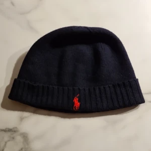 Ralph Lauren Polo mössa svart stl one size. - Polo Ralph Lauren mössa i svart med rött märke, inköpt på Zalando för 639:-. Säljer för att den är för liten, använd 1 gång. Fläckfritt,  djurfritt o rökfritt hem. 
