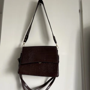 Brun väska - Jättesnygg brun väska med två axelband, ett bredare kort och ett justerbart smalt. Tre innerfack och ett fack på utsidan. Köpt second hand. Mått: 24x19 cm. 