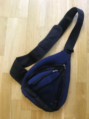 Större midjeväska  - Väska som kan fungera som midjeväska eller på ryggen, i något större modell med flera fack för förvaring och detaljer. På framsidan knäppa väska med kardborre, där även ett litet fack finns för förvaring (se bild 2). Bra skick! 