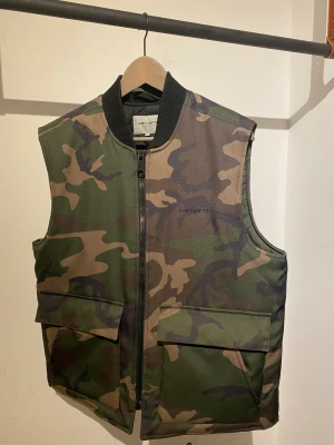 Carhartt camouflage väst - Säljer en Carhartt camouflage väst i storlek M passar som L. Inga synliga fel på den. Inte använd så ofta och är så gott som ny i kvalitén. Man blir väldigt varm i den då den har isolering vilket passar perfekt för höst- eller vintervädret. 