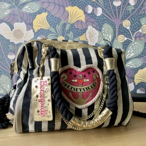 Betsey Johnson Handväska - Väska från Betsey Johnson i marint tema. Svart och vit-randig med guldiga och rosa detaljer med hjärtan, kedjor och båtrattar. Axelremmarna är svarta rep. En guldig tag där det står Betsey Johnson som går att ta av. 