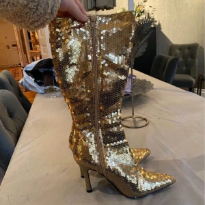 Guldiga stövlar - Ett par jättecoola guldiga paljett stövlar med klack som är perfekta för nyår, halloween, fester, eller andra event. De är använda 2 gånger och är därför så gott som nya. De har även padding på insidan vilket stövlarna mycket skönare att använda. 