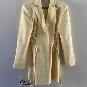 Kavajklänning från Hanna Schönberg - Kavajklänning från Hanna Schönberg i storlek 34, passar 36. Ljusgul och har endast använt en gång på bröllop, bra skick. Sträcker sig till mitten på låren(beroende på längd). Dubbelknäppt och band på sidan som går att ta bort. Jättefin på. Nypris 699 kr