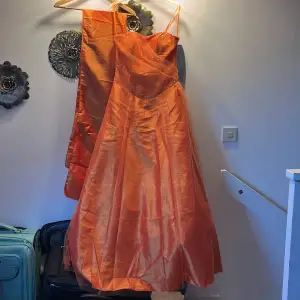 Orange balklänning som har en korsettliknande överdel. Har även en extra sjal som är cirka 2 m i längd och 50 cm i bredd. Finns extra tyg undertill som gör den  puffig men det är enkelt att ta bort ifall man önskar. Skriv vid behov av fler bilder☺️