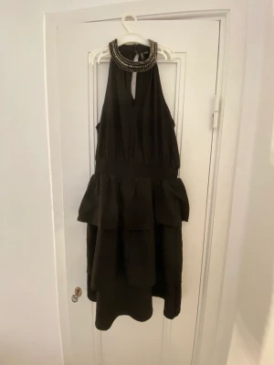 Klänning - Svart klänning från vera moda. Klänningen har en krage med små pärlor som detaljer och sen även flera ”lager” på kjolen som bildar volangerna. Klänningen är använd en gång