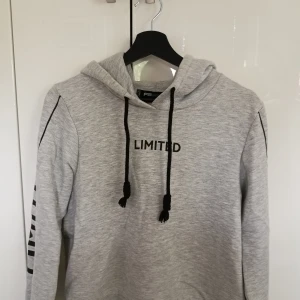 Hoodie med tryck - En grå hoodie med svart text och svarta snören. Texten "Limited" förekommer på framsidan av tröjan samt på ärmarna. Det står att storleken är L, men den är mer som S eller M. Tröjan är från New Yorker. Stussvidd 46cm, längd 46,5cm, Ärmlängd 60 cm.