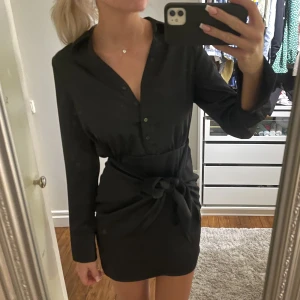 Superfin festklänning (oanvänd, lapp kvar) - Fest/lite mer uppklädd klänning. Svart i siden. Går att stänga och öppna alla knappar så urringningen kan anpassas. Fin smickrande knytdetalj. Säljer endast för den är lite kortare än jag vill (är 179 cm). Helt oanvänd med prislapp kvar, nypris 500kr!