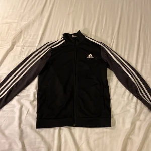 Svart Adidas tröja - Det är en svart Adidas tröja som har en dragkedja som går hela vägen på tröjan storleken är 152 och den har bra skick och inga andra utmärkelser och den har två fickor på utsidan och två på innsidan priset är 150 kr om du vill veta nåt mer så skriv då 