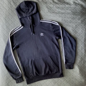 Adidas hoodie - Svart Adidas huvtröja med vita ränder längs ärmarna och logga på framsidan. Helsvart på baksidan. Tvättad några gånger men den är i fint skick.   Längd 67cm, bredd i axlarna 47cm, ärmarna 60cm från axlarna.