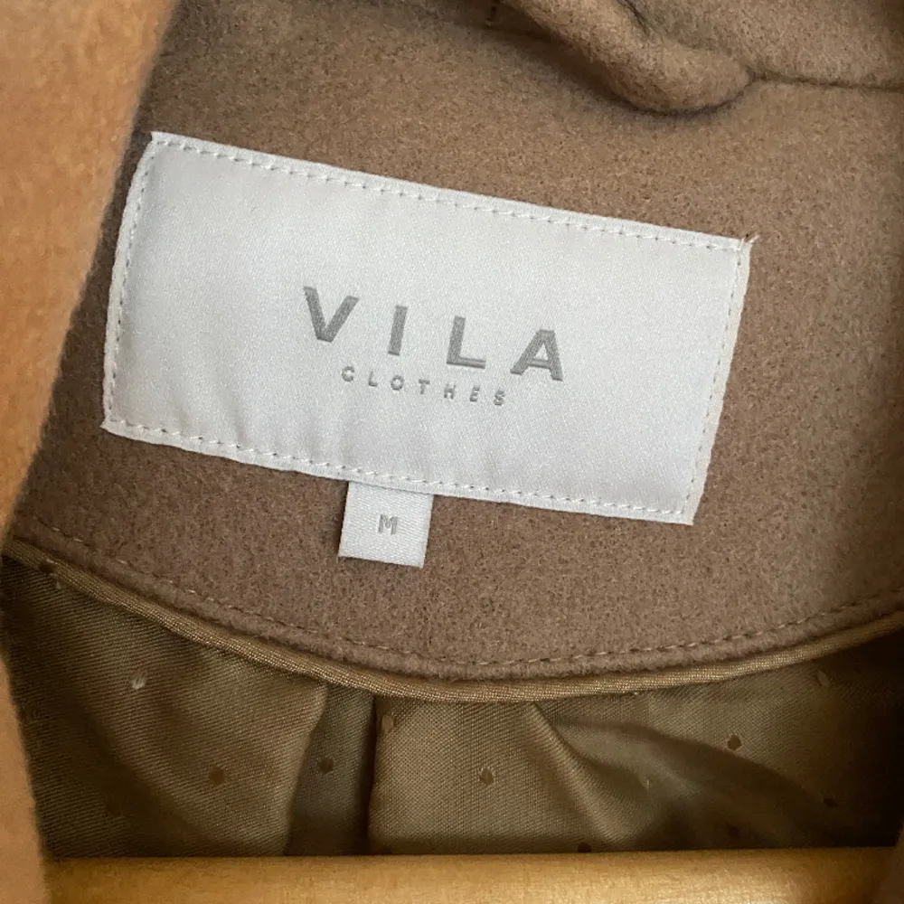 En  beige/ brun kappa ifrån Märket Vila med 2 knappar och ett skärp. Använd 1 gång. . Jackor.