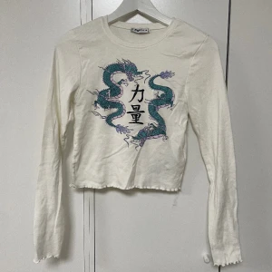 Lång ärmad topp - kawaii - Långärmad topp, vit med intryck av dragon (se bild). På änderna och längst nere av toppen finns det volanger. Den är endast använd 1-2 gånger. Inga tecken på användning. Nypris: 150kr säljer för 89kr