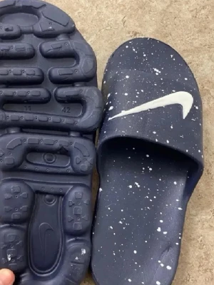 Mörkblå Nike tofflor med vit logga - Säljer ett par mörkblåa Nike tofflor med vit swoosh-logga och vita stänk över hela ovandelen. Tofflorna har en unik, mönstrad sula och är tillverkade i ett mjukt syntetmaterial. Perfekta för att chilla hemma eller till och från träningen.