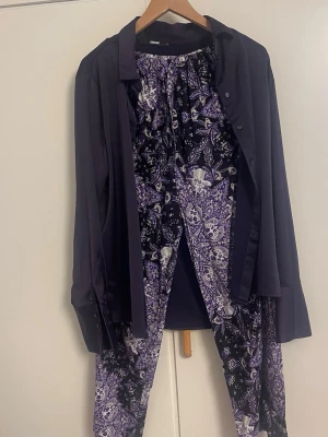 Lila och svart pyjamas-set från Bik Bok - Snyggt pyjamas-set från Bik Bok med långärmad skjorta i enfärgat mörklila och matchande byxor i lila, svart och vitt paisleymönster. Mjukt och skönt material, perfekt för mysiga kvällar hemma.kan passa det M/L