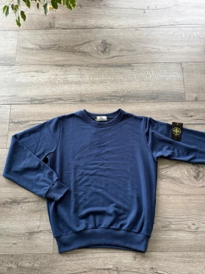  Stone Island Tröja - Säljer en marinblå sweatshirt från Stone Island med klassisk rund hals och ribbade muddar. Tröjan har lång ärm och den ikoniska Stone Island-loggan på vänster ärm. Perfekt för en avslappnad och stilren look.