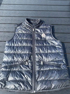 Svart dunväst från Moncler - Snygg svart dunväst från Moncler med glansig finish och ståkrage. Västen har två dragkedjeförsedda fickor framtill och Moncler-logga på bröstet. Perfekt att slänga över en hoodie eller tröja för extra värme. Klassisk quiltad design och lätt vaddering.